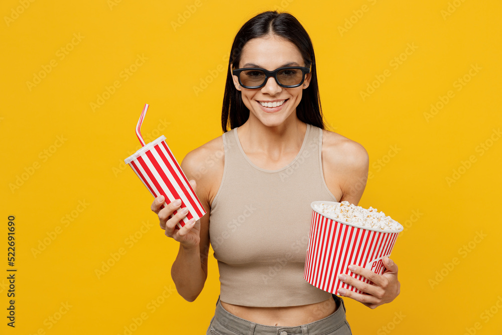 Young smiling cheerful happy fun cool woman she 30s in 3d glasses watch movie film holding bucket of popcorn cup of soda pop in cinema look camera isolated on yellow color background studio portrait.