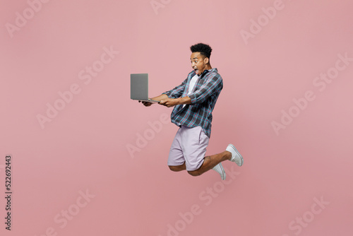 Full body side view young man of African American ethnicity 20s wear blue shirt jump high hold use work on laptop pc computer isolated on plain pastel light pink background. People lifestyle concept.