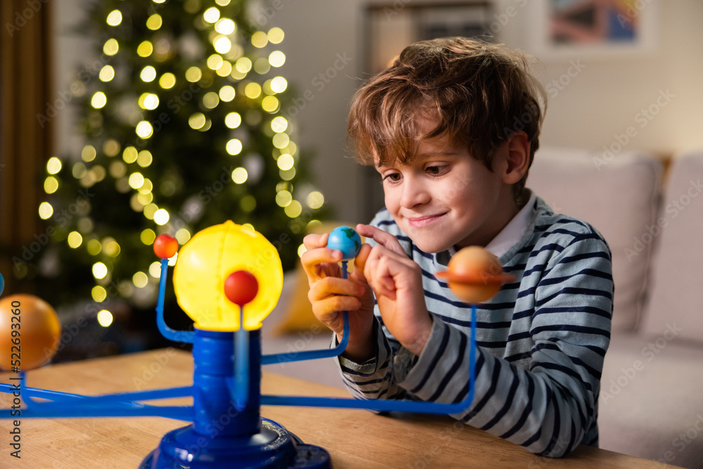 A child plays by a Christmas tree lit with lights, a school-age boy looks at a toy planetary system he built himself, fun combined with learning, the kid holds the planet A child plays by a Christmas tree lit with lights, a school-age boy looks at a toy planetary system he built himself, fun combined with learning, the kid holds the planet
