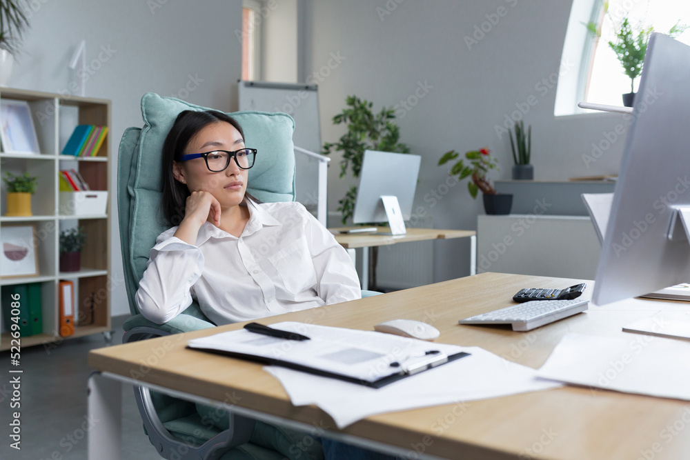 Pensive and sad young Asian businesswoman sitting in the office at the desk in a big chair, thinking about solving a problem Pensive and sad young Asian businesswoman sitting in the office at the desk in a big chair, thinking about solving a problem