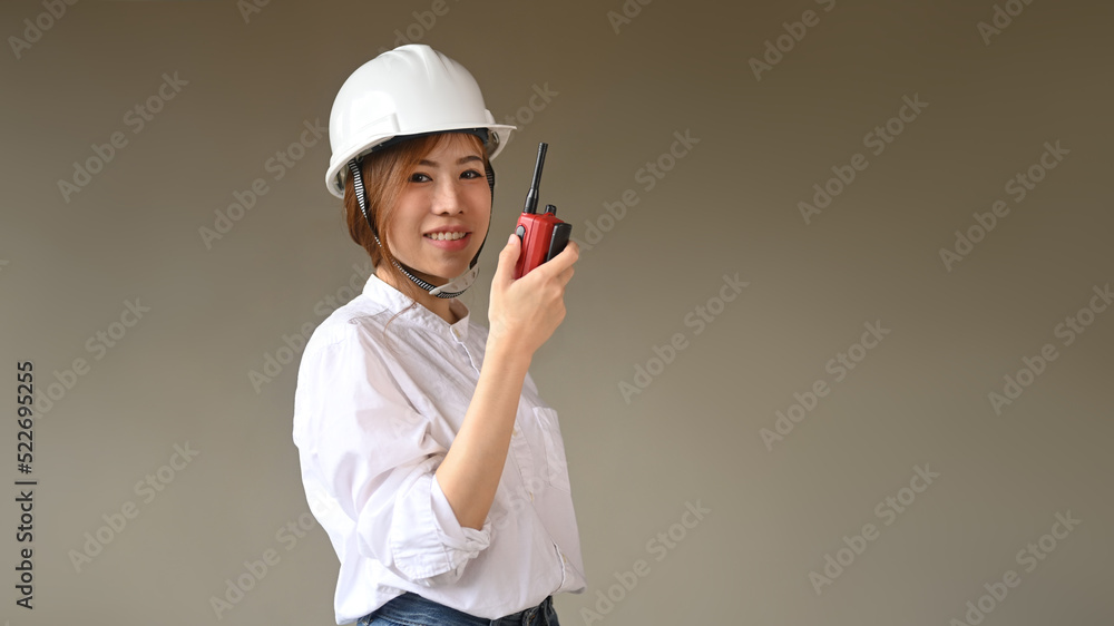 Portrait of female engineer in a protective helmet standing against grey background and looking at camera