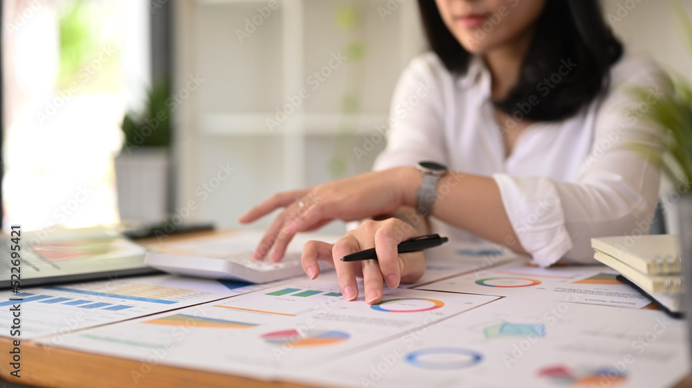 Fototapeta premium Cropped view of female accountant using calculator and examining the numerical data on financial document