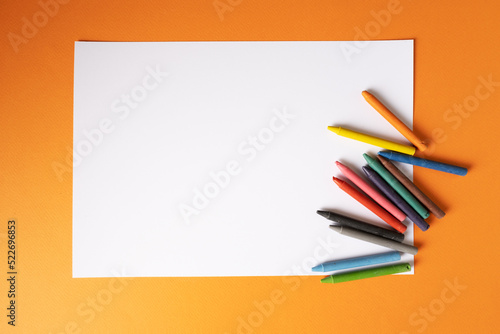 A blank white sheet of paper with wax crayons lies on a orange background