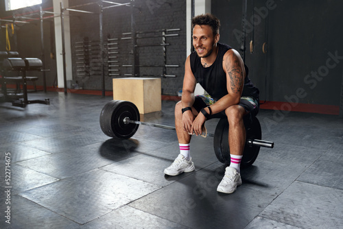 Little break. Handsome Young Man in Sportswear Looking Away with Smile and Relaxing while Sitting at Gym at the Dumbbell. Sport and Recreation Concept 