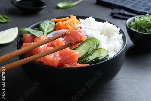 Wooden chopsticks with piece of salmon over delicious poke bowl on grey table, closeup