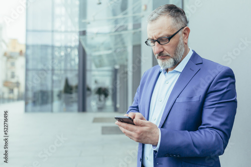 Serious and pensive senior gray-haired man outside office building looking at smartphone screen reading news, businessman in glasses and business suit thinking about decision holding phone in hands