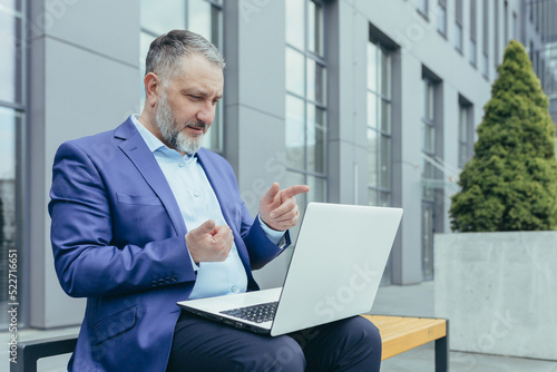 Senior gray-haired successful businessman from office building sitting on bench and using laptop for video call, man in business suit talking remotely, online meeting