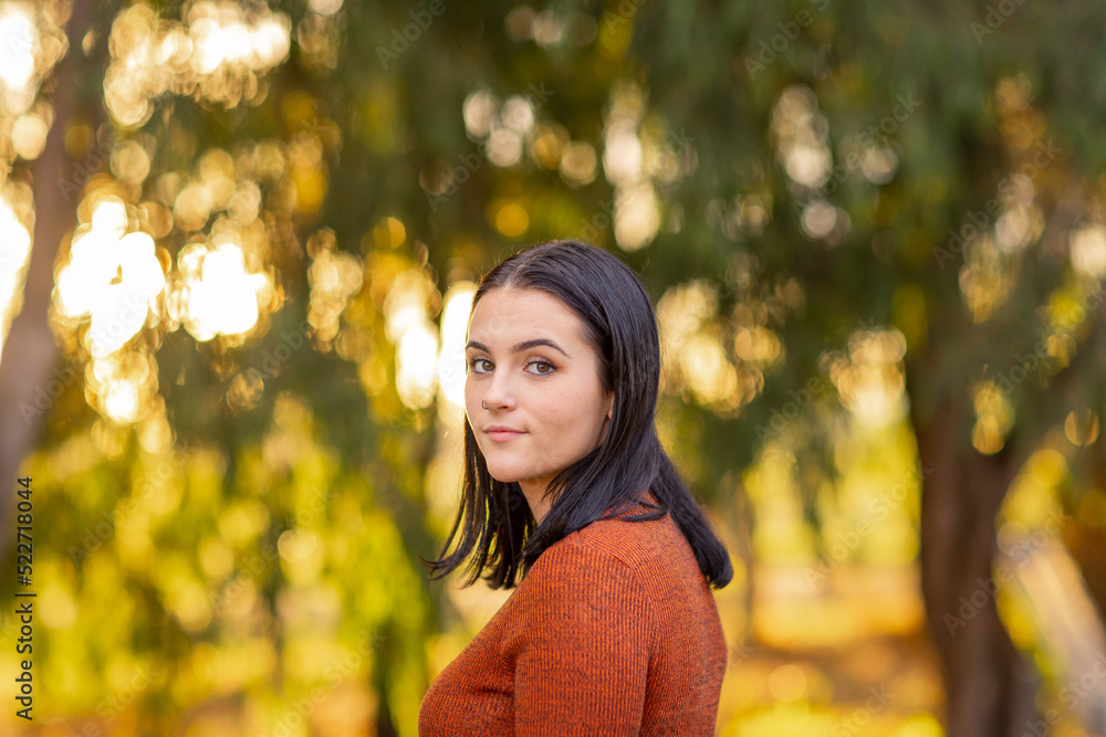 dark-haired teen girl looking over her shoulder against blurry background of trees dark-haired teen girl looking over her shoulder against blurry background of trees