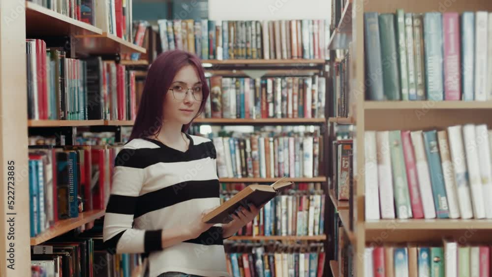 Young free Caucasian woman in glasses with purple hair stands in the library with a book and looks at the camera. Self-education library Young free Caucasian woman in glasses with purple hair stands in the library with a book and looks at the camera. Self-education library