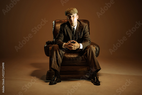 Cinematic portrait of young man, retro 1920s style english gangster wearing suit and cap isolated over dark vintage background. Business, family, art, fashion