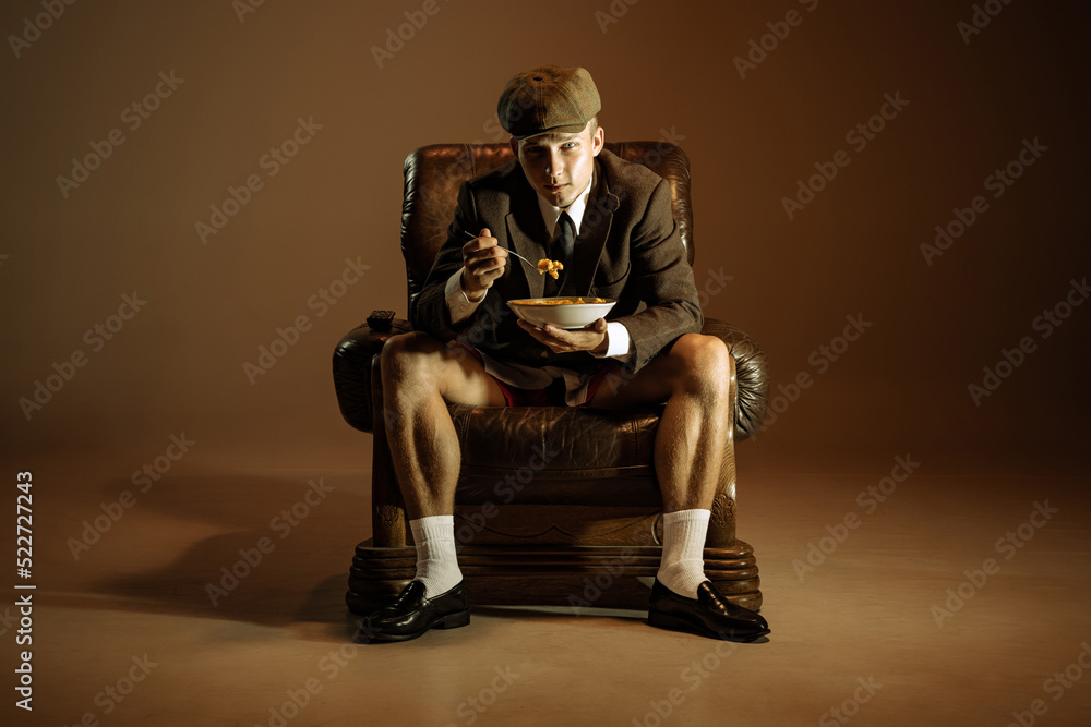 Young man, retro 1920s style english gangster wearing suit and cap tasting pasta isolated over dark vintage background. Business, family, art, fashion Young man, retro 1920s style english gangster wearing suit and cap tasting pasta isolated over dark vintage background. Business, family, art, fashion
