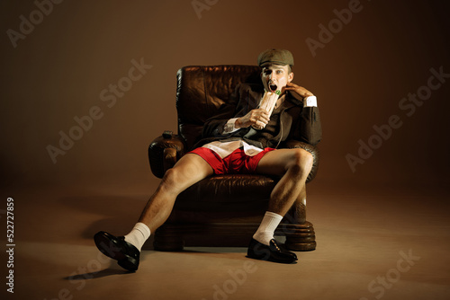 Young man, retro 1920s style english gangster jacket, shorts and cap tasting fast food isolated over dark vintage background. Business, family, art, fashion