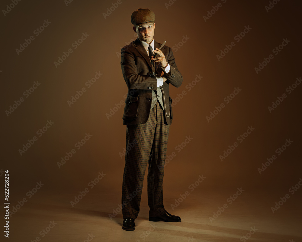Retro style portrait of young man in image of english gangster, businessman wearing suit and cap standing isolated over dark vintage background.
