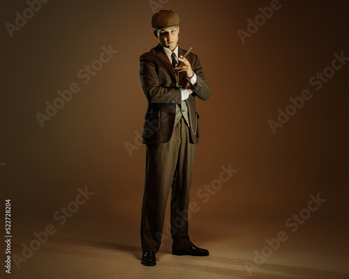 Retro style portrait of young man in image of english gangster, businessman wearing suit and cap standing isolated over dark vintage background.