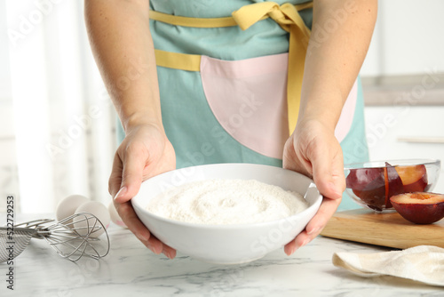 Woman holding bowl with flour at white marble table, closeup. Cooking of delicious plum cake