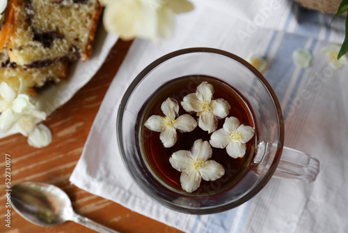 Glass cup of aromatic tea with beautiful jasmine flowers and dessert on wooden table, flat lay
