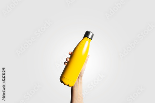 Close-up of female hand holding eco reusable thermo water metal bottle of yellow color on light grey background.