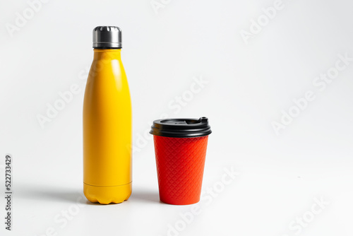 Close-up of metal thermo water bottle of yellow near paper cup for coffee takeaway of red color, on white studio background.