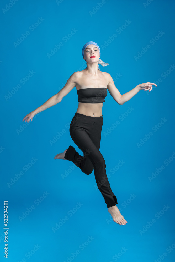 Sport, fashion and make-up concept. Slim and beautiful woman with black and tight sport outfit, scarf and red lipstick jumping in air. Graceful pose freeze in motion. Blue studio background