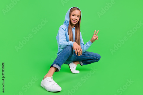 Full length photo of charming little girl show v-sign symbol crouch posing wear stylish blue sportswear isolated on green color background