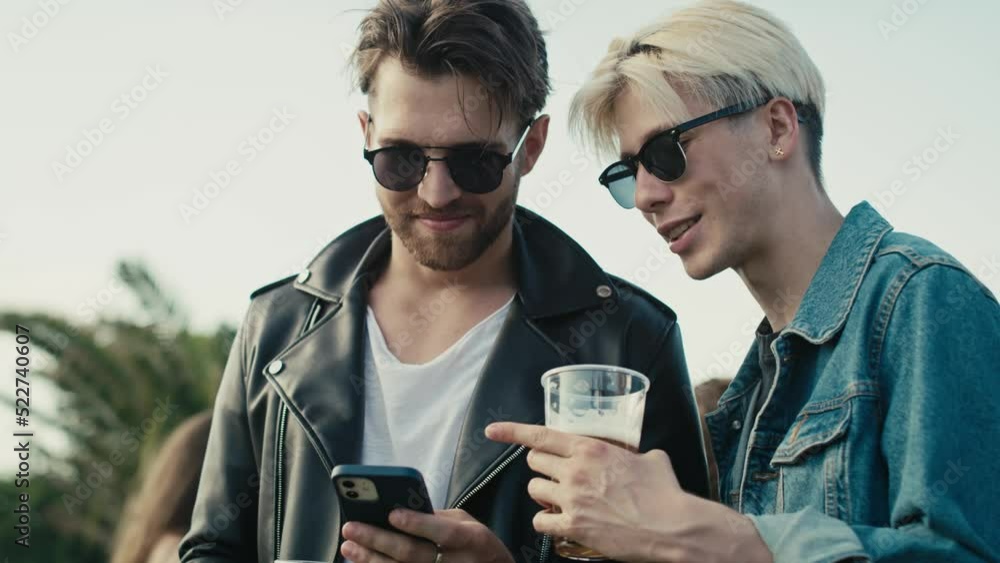 Two young caucasian men browsing phone and having fun on music festival while drinking beer. Shot with RED helium camera in 8K. Two young caucasian men browsing phone and having fun on music festival while drinking beer. Shot with RED helium camera in 8K.