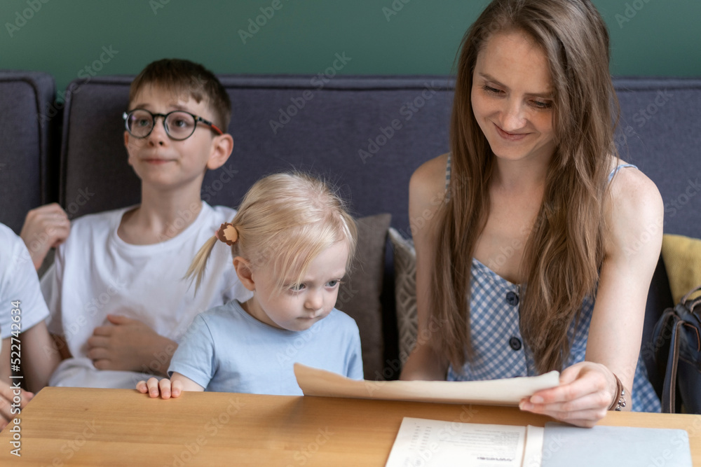 Beautiful young mother relaxes in cafe with her children reading the menu and smiling. Large family relaxes in cafe. Beautiful young mother relaxes in cafe with her children reading the menu and smiling. Large family relaxes in cafe.