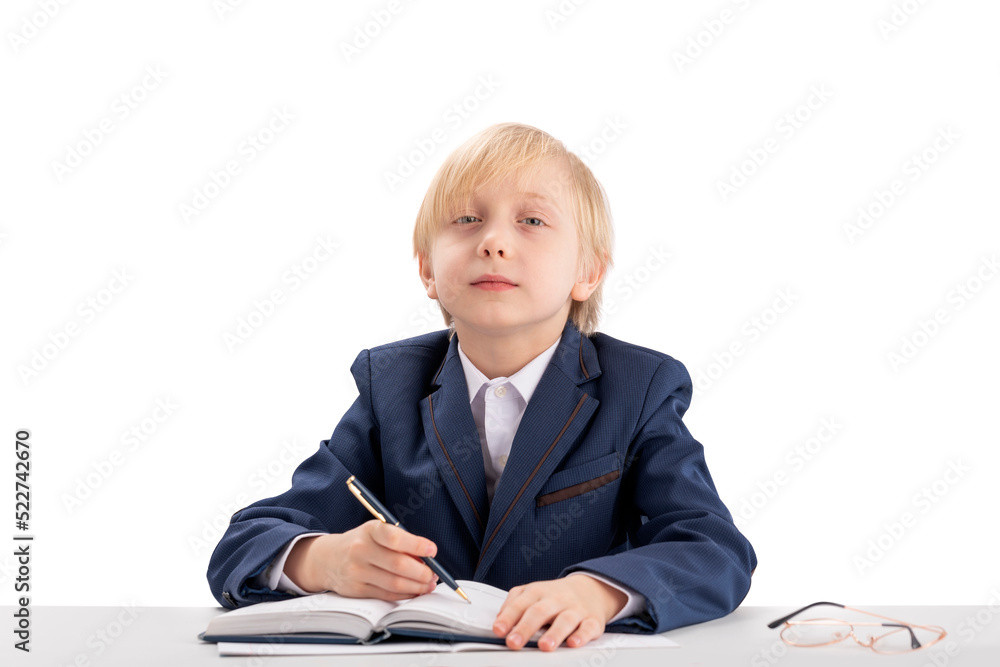 Child like boss sits at table and writes in notebook. Diligent student writes in copybook. Blond boy in jacket and shirt. Child like boss sits at table and writes in notebook. Diligent student writes in copybook. Blond boy in jacket and shirt.