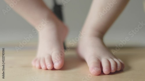 tiny barefoot baby feet of sitting child toes and heels moving on wooden surface. close-up view caucasian legs and feet of infant toddler kid stepping, hanging, touching light wood floor indoors.  photo