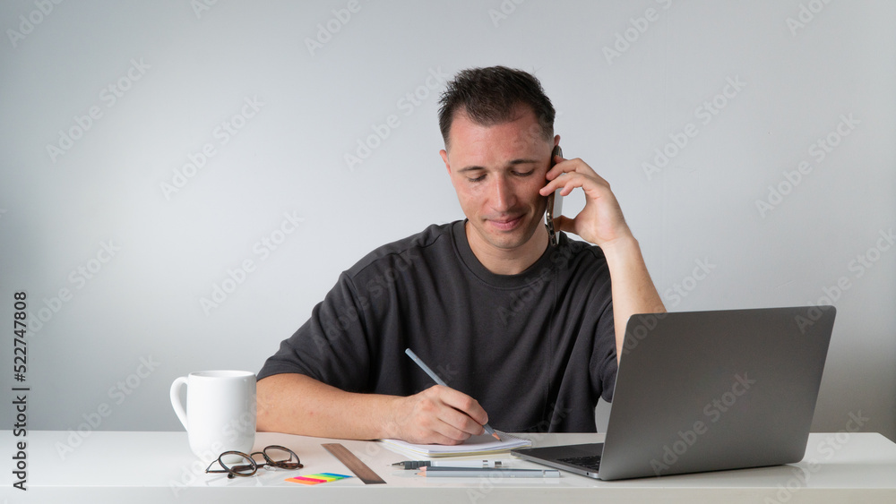 Male student writes a lecture in a notebook and talks on the phone with a laptop Male student writes a lecture in a notebook and talks on the phone with a laptop