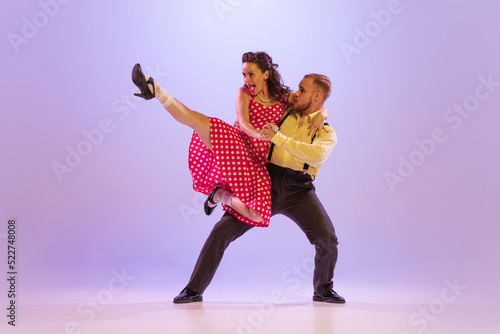 Active and emotional couple in colorful retro style costumes dancing incendiary dances isolated on purple background in neon light. Concept of art, culture photo