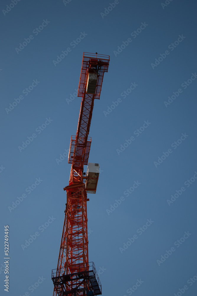 A red orange construction tall crane against a clear blue blue sky in the sunshine of the day in the rays of the sun. Construction site on the background blue sky, panoramic mock-up A red orange construction tall crane against a clear blue blue sky in the sunshine of the day in the rays of the sun. Construction site on the background blue sky, panoramic mock-up