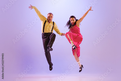 Active and emotional couple in colorful retro style costumes dancing incendiary dances isolated on purple background in neon light. Concept of art, culture