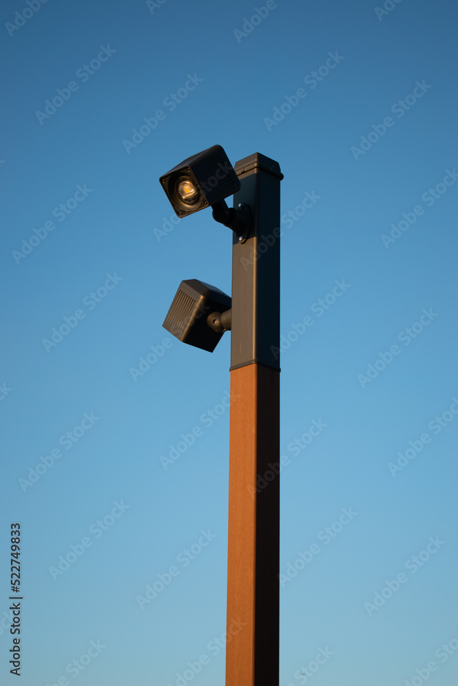 A tall insulated lantern or CCTV camera on a wooden pole or pole with an iron black or gray coating on top in golden sunlight against a clear blue sky A tall insulated lantern or CCTV camera on a wooden pole or pole with an iron black or gray coating on top in golden sunlight against a clear blue sky