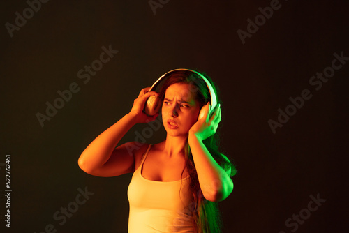 Portrait of emotive young woman in headphones posing over brown background in neon light