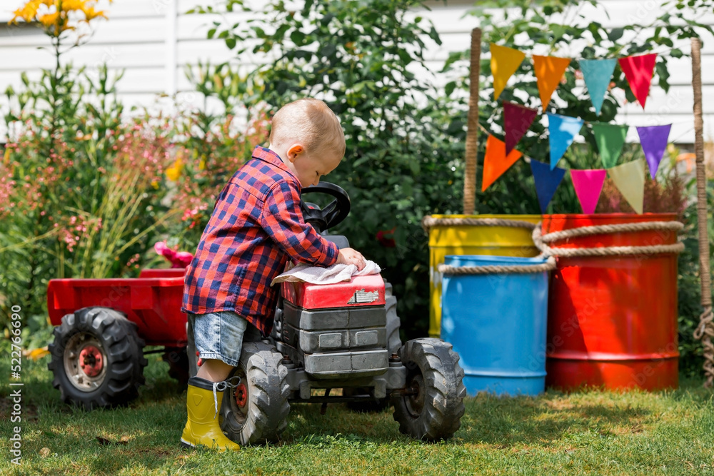 A cheerful boy dressed in a rustic style washes a tractor with a rag in the garden in the village. Playing in nature. The concept of raising a child and teaching them to work. A cheerful boy dressed in a rustic style washes a tractor with a rag in the garden in the village. Playing in nature. The concept of raising a child and teaching them to work.
