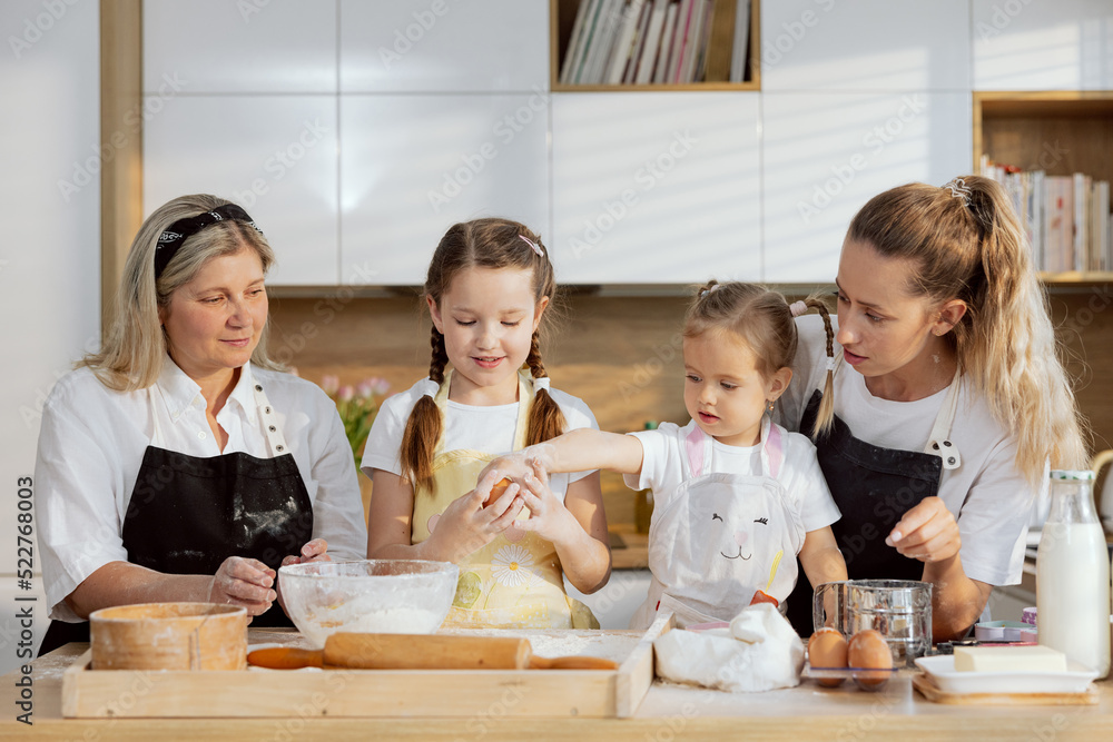 Happy family wearing aprons cooking baking in modern kitchen. Daughters trying beating eggs into large bowl for kneading dough. Baking cooking homemade pizza bread cookies. Happy family wearing aprons cooking baking in modern kitchen. Daughters trying beating eggs into large bowl for kneading dough. Baking cooking homemade pizza bread cookies.