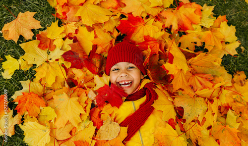 A girl with a wide smile lies on a carpet of red and yellow leaves in an autumn park.