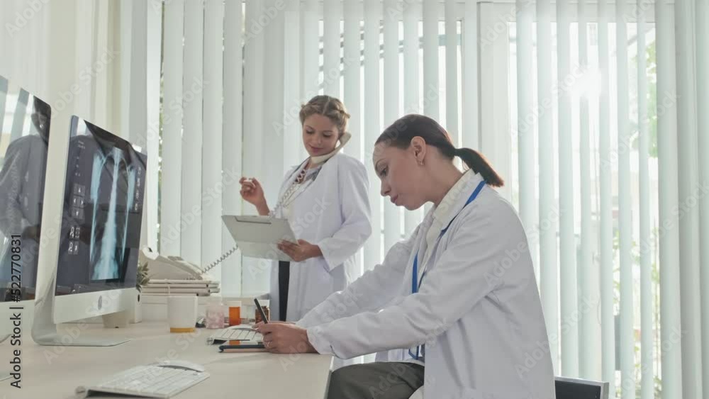 Female doctor examining x ray image on computer and taking notes on clipboard as her colleague talking on phone at work in clinic Female doctor examining x ray image on computer and taking notes on clipboard as her colleague talking on phone at work in clinic