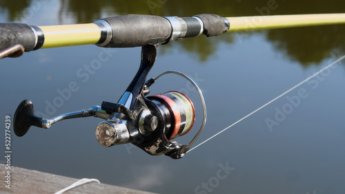 An old shabby spinning rod with a red reel against the blue water in the pond. The concept of outdoor recreation and hobbies.