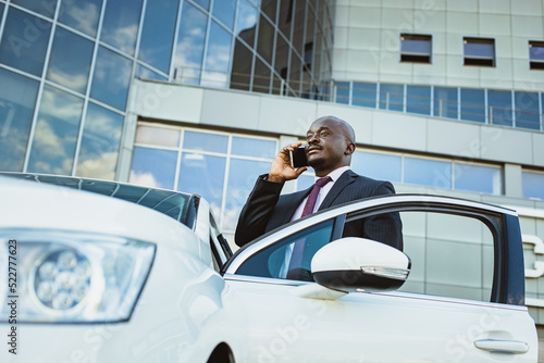 Portrait of a handsome successful African American businessman in a suit stands near to his expensive luxury car and talking by phone