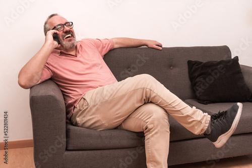 Bearded man sitting on a sofa and laughing happily while talking on the cell phone.