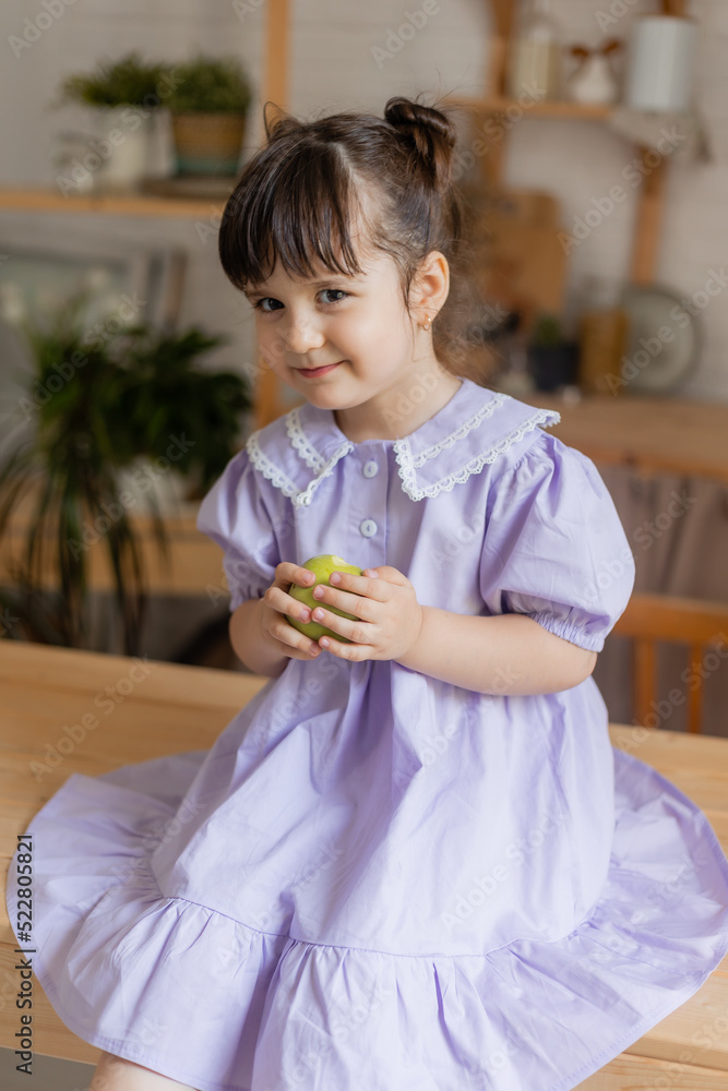 charming little girl in a lilac dress eats a green apple in the kitchen. space for text, banner charming little girl in a lilac dress eats a green apple in the kitchen. space for text, banner