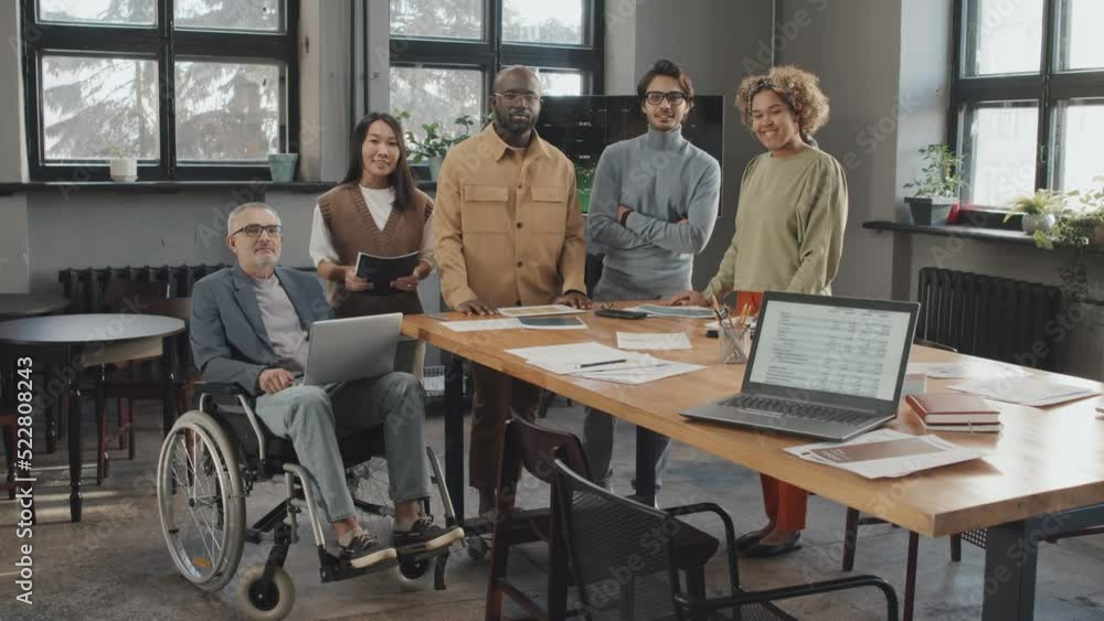 Long portrait of Asian woman, African American girl, Black man, Middle Eastern male colleague and senior Caucasian businessman in wheelchair at conference table looking and smiling on camera Long portrait of Asian woman, African American girl, Black man, Middle Eastern male colleague and senior Caucasian businessman in wheelchair at conference table looking and smiling on camera