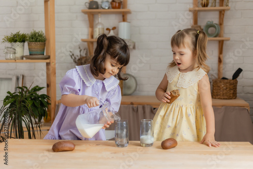 two little girls in the kitchen pour milk from a glass jug into glasses and drink. space for text