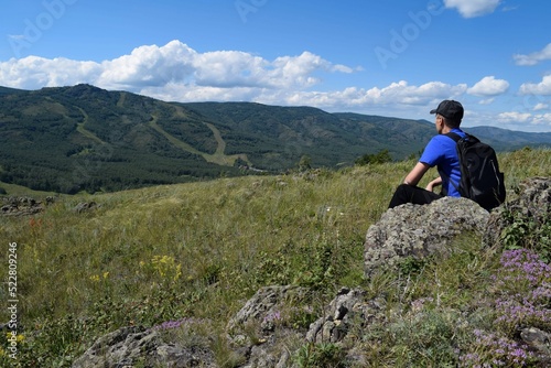 A man with a backpack sits on a rock and looks at the mountain on a summer day