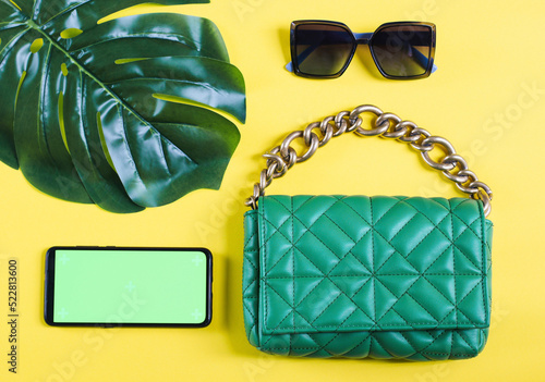 Stylish green quilted bag with big gold chain with sunglasses, manser leaf and green screen smartphone photo