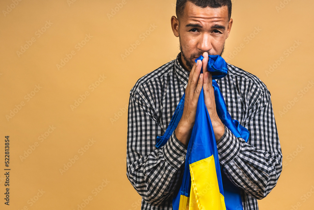 © denis_vermenko - Portrait of sad negative african american black guy holding ukrainian flag, isolated over beige background.