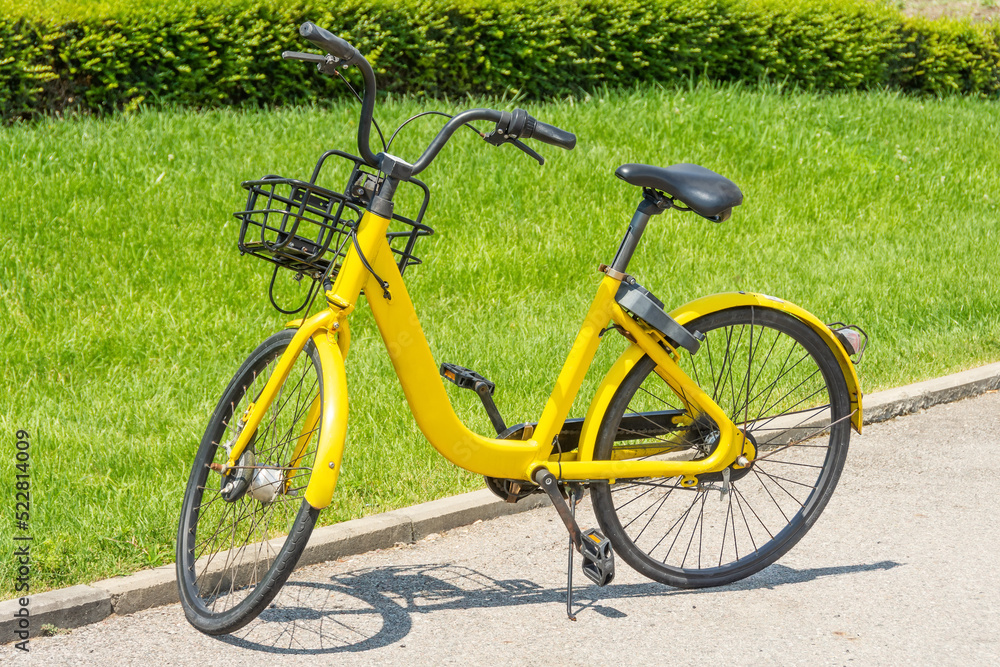 Yellow bicycle with a basket on the footboard in the parking lot near the green city lawn on the footpath.