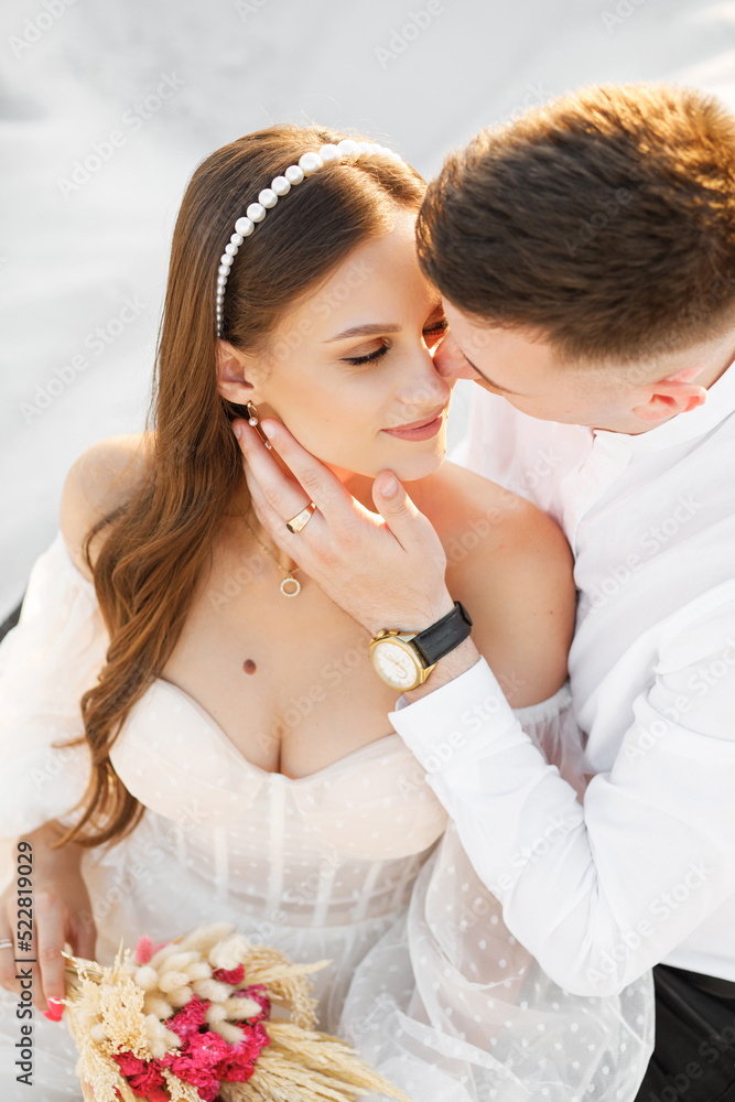 Gentle portrait of the newlyweds on the background of white sand. A man kisses his wife on the cheek. Soft light, natural colors Gentle portrait of the newlyweds on the background of white sand. A man kisses his wife on the cheek. Soft light, natural colors