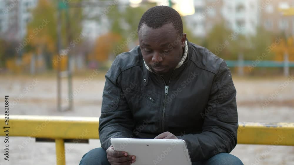 African american man sits at sport ground and works with tablet computer. African uses tablet computer, flips through feed on social networks African american man sits at sport ground and works with tablet computer. African uses tablet computer, flips through feed on social networks